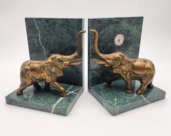 May include: A pair of bookends featuring two brass elephant figures attached to dark green marble L-shaped bases. Each elephant is depicted with its trunk raised upward and tusks extending forward. A small circular sticker is visible on one base.