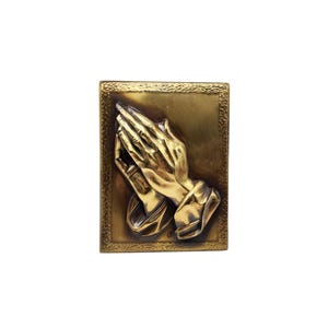 May include: A rectangular, gold-toned relief sculpture featuring clasped hands in a prayerful pose. The hands are detailed with visible fingers and a textured surface, set against a gold background within a decorative frame.
