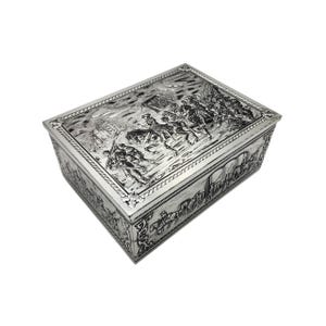 May include: A rectangular silver box with detailed relief carvings depicting a historical battle scene. The box features a hinged lid and intricate border designs, showcasing a vintage aesthetic. The carvings include soldiers on horseback and other figures.