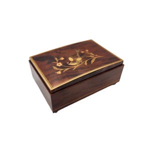 May include: A rectangular wooden jewelry box with a dark brown finish. The lid features an inlaid floral design with gold and light brown accents. A thin gold band frames the lid. The box is a classic decorative item.