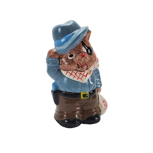 May include: A ceramic figurine of a bear dressed as a cowboy, wearing a blue hat, jacket, and brown pants. The bear has an eye patch, a red and white bandana, and is holding a money bag with a dollar sign.