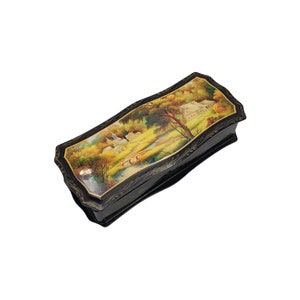 May include: A black lacquered wooden box with a hinged lid. The lid is decorated with a painted scene of a village in a forest setting. The scene is painted in shades of gold, green, and brown.