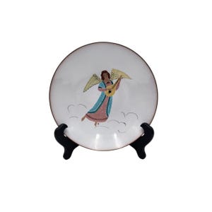 May include: A white decorative plate with a colourful illustration of an angel playing a lute. The angel has brown hair, yellow wings, and wears a blue and pink robe. The plate is displayed on a black stand.