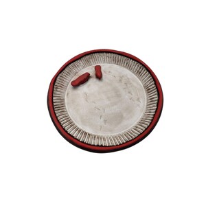 Puede incluir: Un plato de cerámica hecho a mano con un borde rojo y un centro blanco roto texturizado. El plato presenta una serie de líneas verticales alrededor del borde y dos pequeñas formas rojas abstractas en la superficie. El plato es redondo.