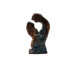 May include: A small, abstract ceramic sculpture in shades of brown and blue. The piece features two intertwined figures, with a rough, textured surface. The sculpture has a base and a curved, open design.