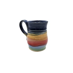Puede incluir: Una taza de cerámica con una sección superior azul oscuro y un asa. El cuerpo inferior presenta bandas horizontales de esmalte amarillo, naranja, rojo, azul claro y verde. La taza tiene una forma redondeada y ligeramente abombada.