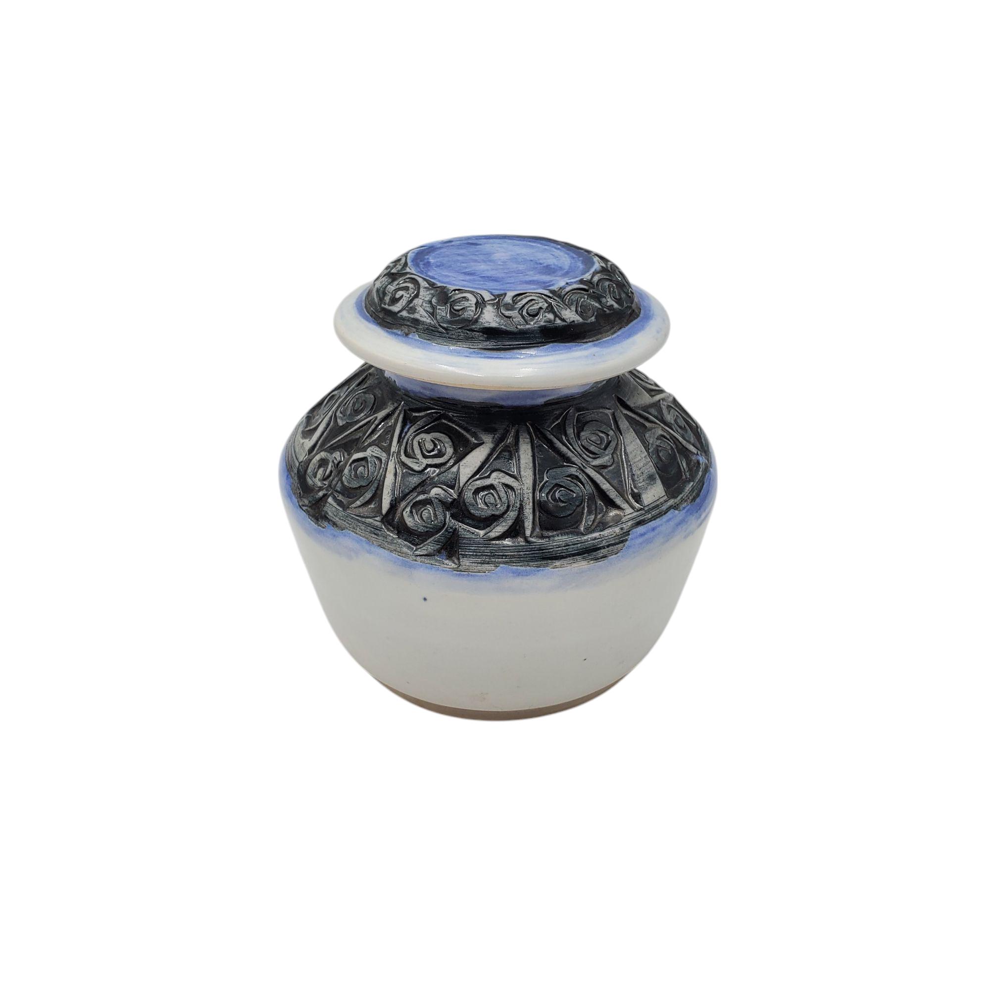 May include: A small white ceramic jar with a blue and black lid. The jar has a black floral design around the middle.