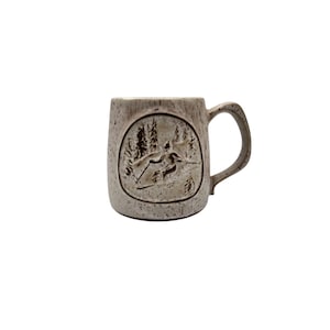 May include: A ceramic mug with a textured, speckled finish. The mug features a circular embossed design of a skier on a mountain slope surrounded by trees. The handle is curved and the mug has a neutral colour palette.