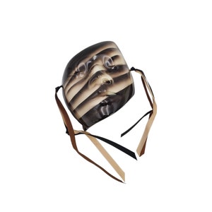 May include: A black and white striped ceramic mask with brown and black ribbon ties. The mask has a human face with closed eyes and a slightly open mouth.