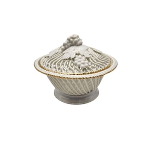 May include: White ceramic lidded basket with a gold trim. The lid features a grape cluster and leaf design. The basket has a woven pattern and sits on a pedestal base. A decorative piece for the home.