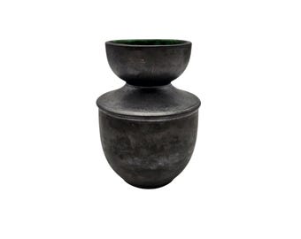 Può includere: Un vaso in ceramica grigio scuro con coperchio. Il vaso ha una base bulbosa e una forma a ciotola più piccola e rovesciata sulla parte superiore. L'interno della ciotola superiore ha una tonalità verde. Il pezzo ha una finitura opaca.