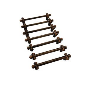May include: A collection of seven bronze-coloured cabinet pulls. Each pull has a cross-shaped design at each end and a textured, diamond-patterned bar in the middle. The pulls are arranged diagonally against a white background.