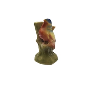 May include: A ceramic bird figurine perched on a tree stump. The bird has a blue head, orange chest, and yellow wings. The tree stump is green and brown. The figurine is a decorative item.