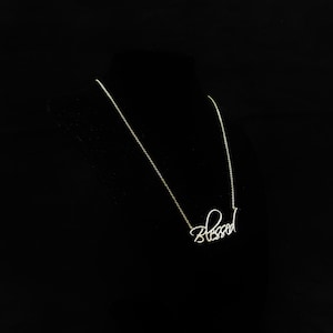 Puede incluir: Un delicado collar plateado con la palabra "Blessed" en escritura cursiva. El colgante cuelga de una fina cadena, sobre un fondo negro. El collar es una pieza de joyería sencilla pero elegante.