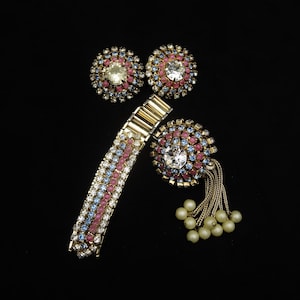 Puede incluir: Un conjunto de joyas vintage con una pulsera dorada y dos pares de pendientes, todos adornados con pedrería rosa, azul y transparente. Un broche a juego con perlas de imitación colgantes completa el conjunto. Las joyas están sobre un fondo negro.