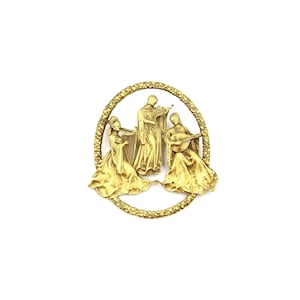 May include: A circular gold-toned brooch featuring three figures in flowing robes playing musical instruments, including a harp, a violin, and a lute. The figures are enclosed within a textured, wreath-like border with a detailed floral pattern.