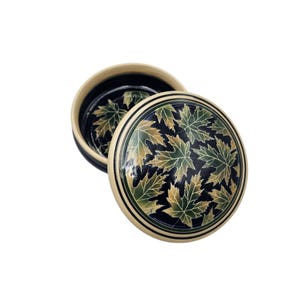 May include: A round, wooden box with a lid. The lid and interior base are black with a pattern of green and gold maple leaves. The rim of the box is cream-colored with a black and green border.
