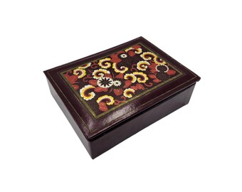 May include: A rectangular, dark brown jewelry box with a decorative floral design on the lid. The lid features a pattern of red, yellow, and white flowers and swirling shapes against a dark background. The box is closed and sits on a white surface.