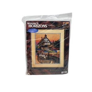 May include: A Monarch Horizons embroidery kit featuring a Longstitch Country Manor scene. The artwork depicts a detailed house with a reflection in the water, framed in a dark brown border. The kit is sealed in a clear plastic bag.