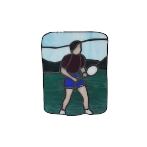 May include: Stained glass artwork depicting a person playing tennis. The person is wearing a maroon shirt, blue shorts, and is holding a white tennis racket. The background is a blue sky with white clouds and green grass.