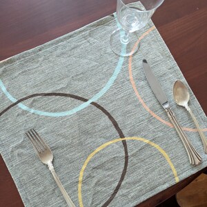 May include: A gray placemat with overlapping circles in brown, yellow, light blue, and peach. Silverware, including a fork, knife, and spoon, is arranged on the placemat. A clear wine glass sits on the placemat.