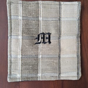 May include: Square, beige and grey plaid cloth napkin with a black embroidered letter "M" in the centre. The napkin has a stitched border and a woven texture. The napkin is likely used for dining.