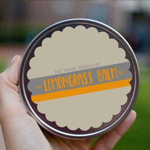 May include: A silver tin with a cream-coloured label that says "THE HOME HERBALIST LEMONGRASS BALM" in black and orange lettering.