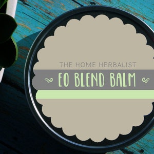 May include: A round metal tin with a scalloped edge and a label that reads "THE HOME HERBALIST EO BLEND BALM". The label is gray with a green stripe and white text.
