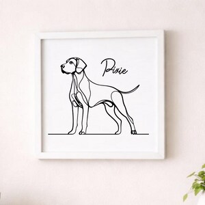 May include: A framed minimalist line art print of a dog in black ink. The dog is standing in profile, with the name "Pixie" written above it in cursive. The artwork is displayed on a white wall above a wooden cabinet.