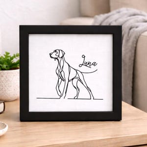 Puede incluir: Un marco negro con un dibujo lineal minimalista de un perro. El perro está representado de pie con el nombre "Luna" en cursiva. La obra de arte se exhibe sobre una superficie de madera.