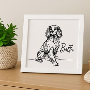 May include: A framed black and white line drawing of a dog, with the name "Bella" in cursive. The artwork is in a white square frame, placed on a wooden surface. A small potted plant is visible on the left.