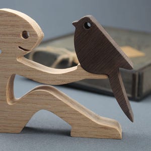 May include: Wooden sculpture of a figure with a bird. The figure is light brown with a smiling face and is holding a dark brown bird on its arm. The bird has a circular eye. The sculpture is on a gray surface.