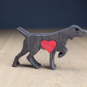 May include: A wooden dog figurine in a walking pose, painted dark grey with a red heart-shaped cutout on its side. The dog is made of wood and has a smooth finish. The figurine is displayed on a light-coloured wooden surface.