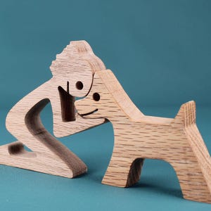 May include: Wooden sculpture depicting a person kneeling to pet a dog. The figures are crafted from light-colored wood, with simple, rounded features and visible wood grain. The dog has a small tail and a smiling face.