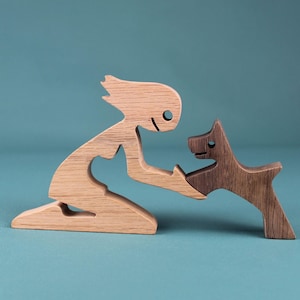 May include: A wooden figurine of a person kneeling and reaching out to a wooden dog figurine. The person is made of light wood and the dog is made of dark wood.