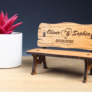 Könnte beinhalten: Eine kleine Holzbank mit einem Herz und den Namen Oliver & Sophia, die auf die Rückenlehne graviert sind. Das Datum 25/05/2025 ist ebenfalls auf die Rückenlehne graviert.