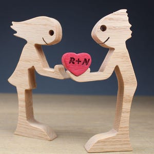 May include: Wooden figurine of two stylized figures holding a red heart with the letters "R+N". The figures are light brown with simple facial features. The heart is a vibrant red, creating a focal point.