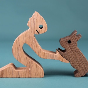 May include: Wooden sculpture of a person kneeling and interacting with a dog. The person is light-colored wood, while the dog is a darker wood. The sculpture is set against a teal background.