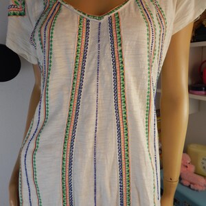May include: White short-sleeve shirt with a V-neck and colorful embroidered details. The embroidery features vertical stripes with blue, green, and coral accents. The shirt is made of a soft, lightweight fabric.