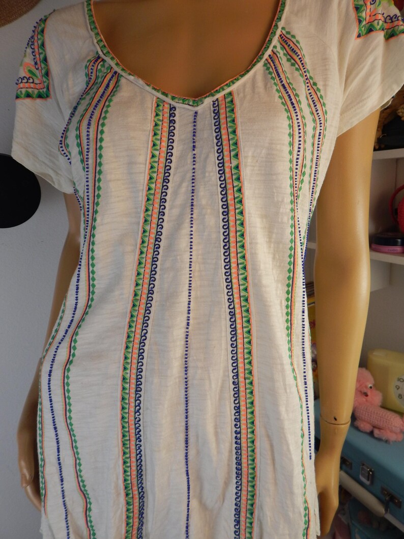 May include: White short-sleeve shirt with a V-neck and colorful embroidered details. The embroidery features vertical stripes with blue, green, and coral accents. The shirt is made of a soft, lightweight fabric.
