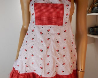 May include: A white apron with a red strawberry pattern and a red polka dot pocket. The apron has a red ruffle at the bottom and red halter straps. The apron is designed for kitchen use.