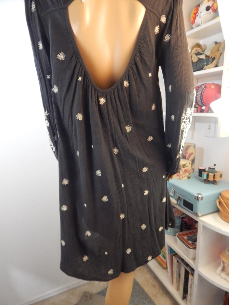 May include: A dark gray dress with long sleeves and a low back. The dress is adorned with small, white floral embroidery scattered across the fabric. The sleeves also feature white embroidery near the cuffs. The dress has a loose, flowing silhouette.