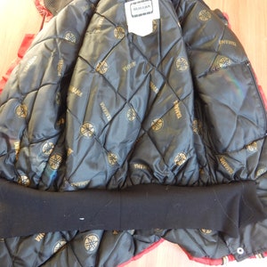May include: Red and black Yamaha jacket. The inside lining is black with a quilted pattern and gold Yamaha logos. The jacket has a black ribbed collar and cuffs. A white tag with the Yamaha logo is visible.