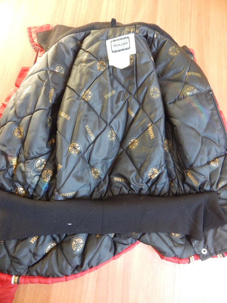 May include: Red and black Yamaha jacket. The inside lining is black with a quilted pattern and gold Yamaha logos. The jacket has a black ribbed collar and cuffs. A white tag with the Yamaha logo is visible.