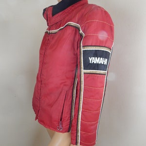 May include: A red Yamaha racing jacket with black and gold accents. The jacket features a black collar, a zipper closure, and the Yamaha logo on the sleeve. The jacket has white and gold stripes down the sleeves.