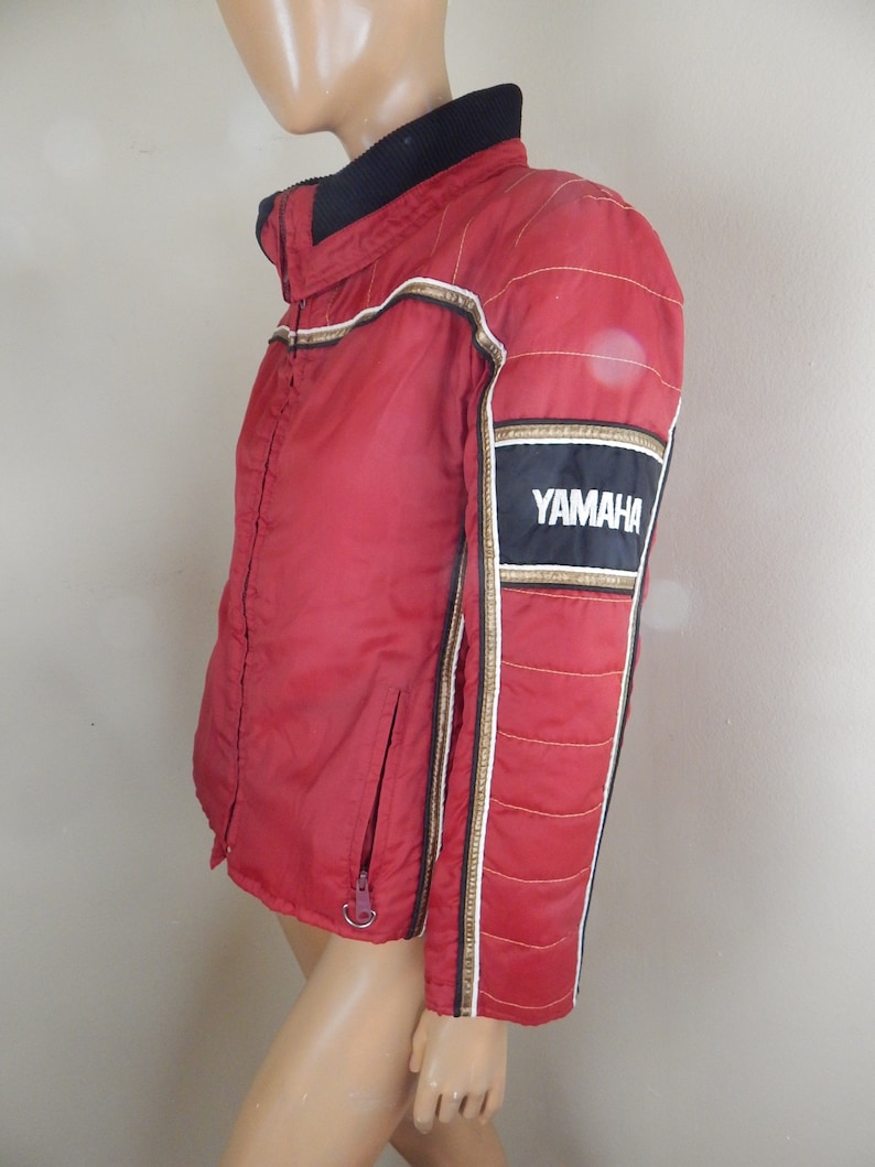 May include: A red Yamaha racing jacket with black and gold accents. The jacket features a black collar, a zipper closure, and the Yamaha logo on the sleeve. The jacket has white and gold stripes down the sleeves.