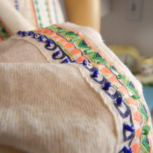 May include: Close-up of a cream-colored garment with decorative trim. The trim features a pattern of orange, green, and blue accents, including small blue beads. The fabric appears soft and textured.