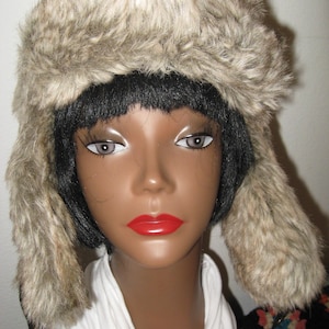 May include: A faux fur trapper hat in shades of brown and tan, with ear flaps. The hat is displayed on a mannequin head with black hair, red lipstick, and a white shirt. This winter hat is designed to keep you warm.