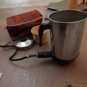 May include: A vintage electric coffee pot with a black handle and power cord. A red plaid travel case, a small beige cup, and a silver lid are also present. The items are on a light brown surface.