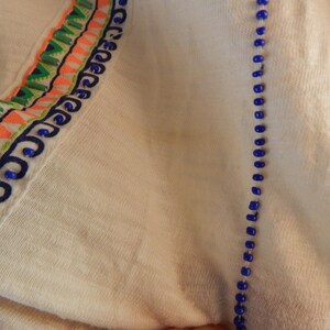 May include: Close-up of a cream-colored fabric with decorative embroidery. The embroidery features a combination of green, orange, and blue colors. A line of blue beads runs vertically along the fabric's edge, adding a touch of elegance.